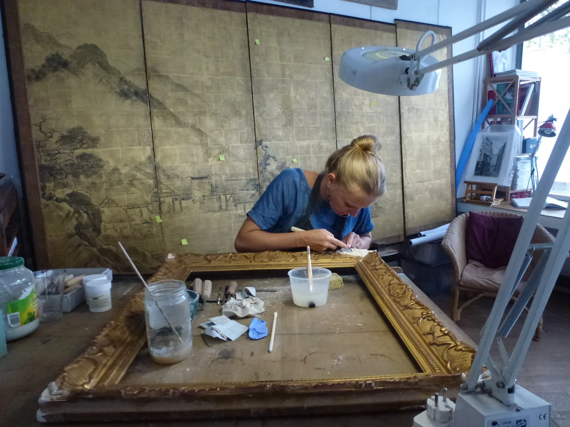 Sibille Gempp restaure un cadre doré à l'atelier, devant un paravent japonais
